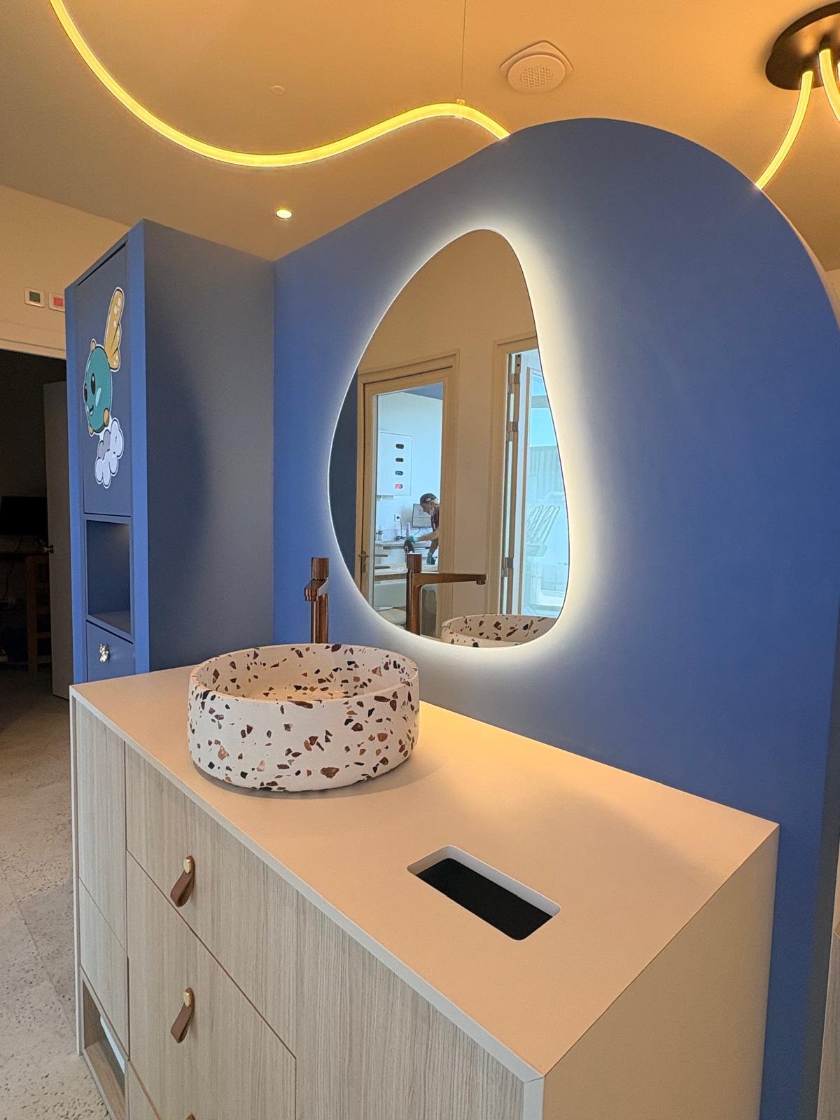 Sanitaires enfants — miroir organique rétro-éclairé, vasque terrazzo et meuble bois sur fond bleu