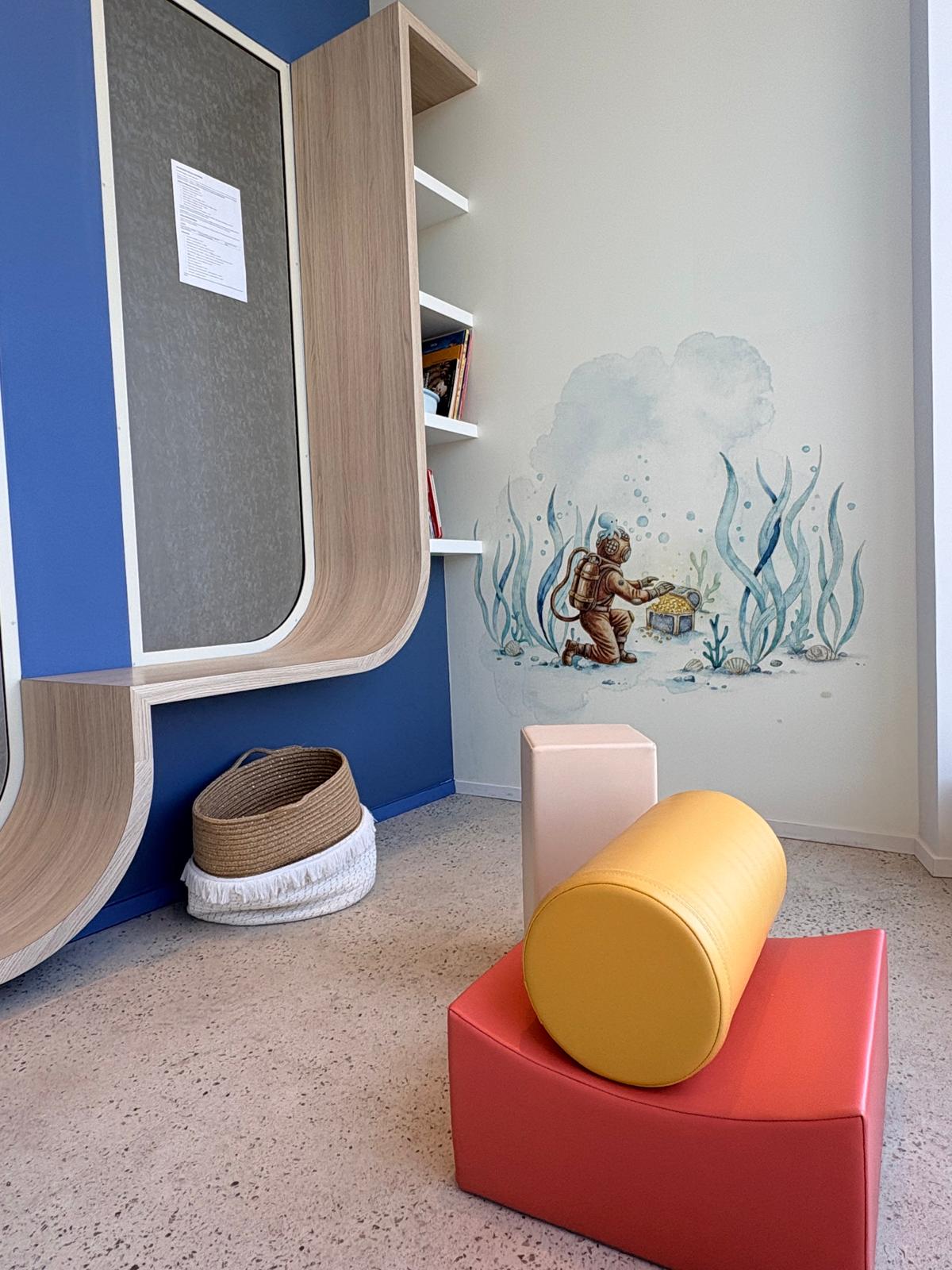 Coin enfants — assises mousse colorées, bibliothèque cintrée bois et bleu, fresque scaphandrier